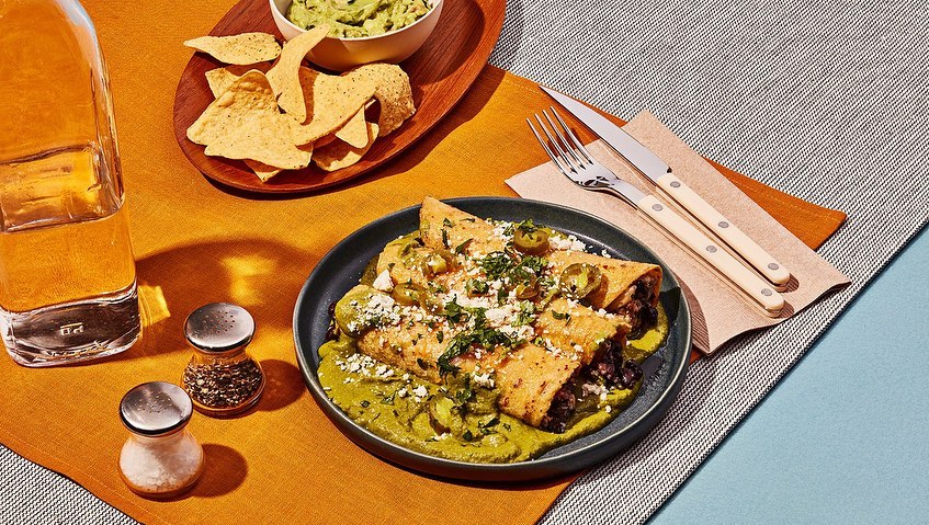 Overhead shot of enchiladas covered in green sauce and cheese on a plate, accompanied by a drink, chips, and guacamole.