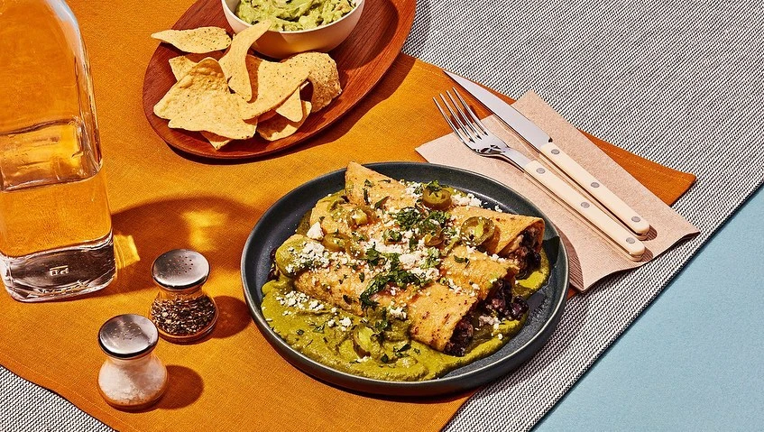 Overhead shot of enchiladas covered in green sauce and cheese on a plate, accompanied by a drink, chips, and guacamole.