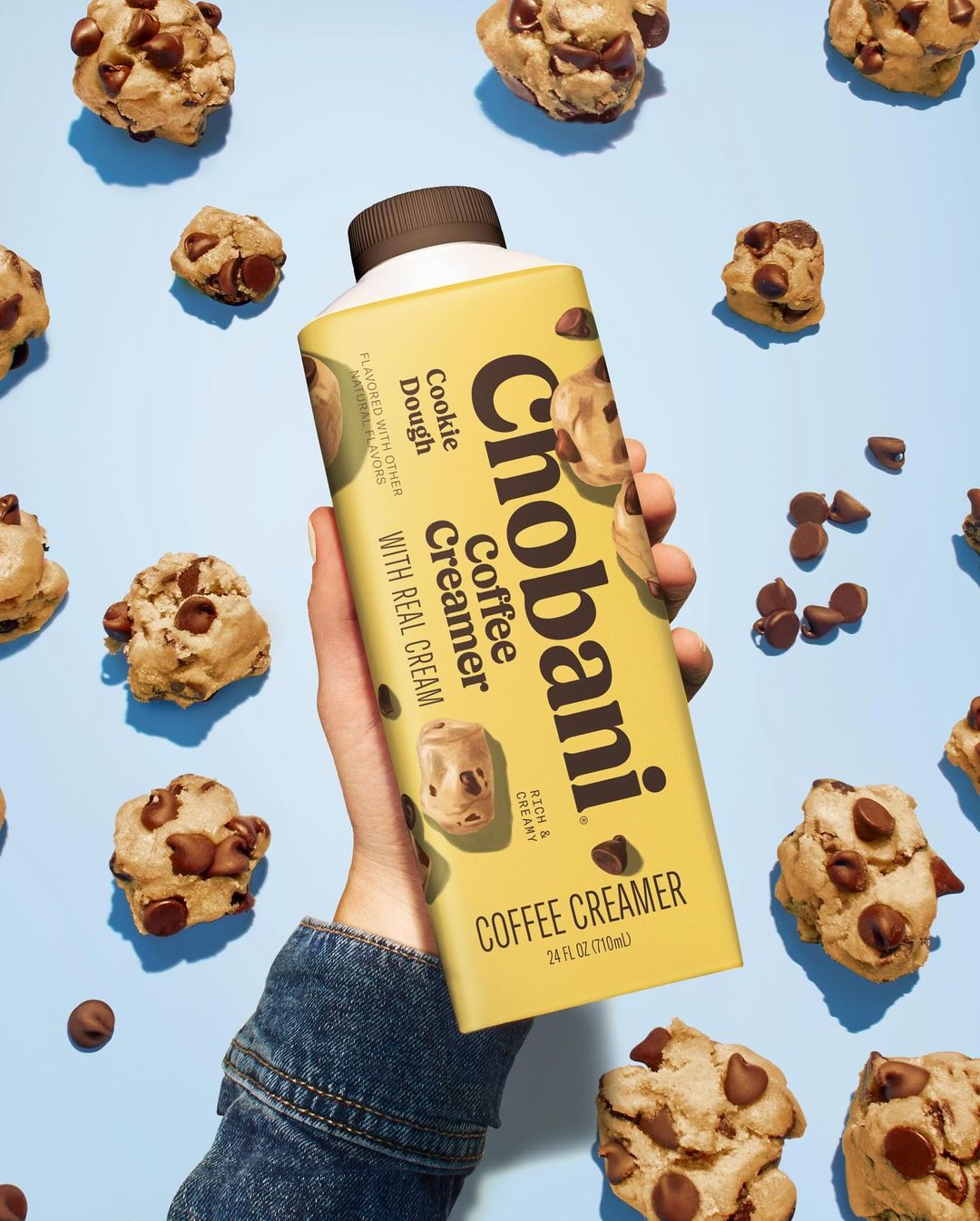 Hand holds Chobani cookie dough coffee creamer with scattered cookie dough pieces on a light blue background in a studio shot.