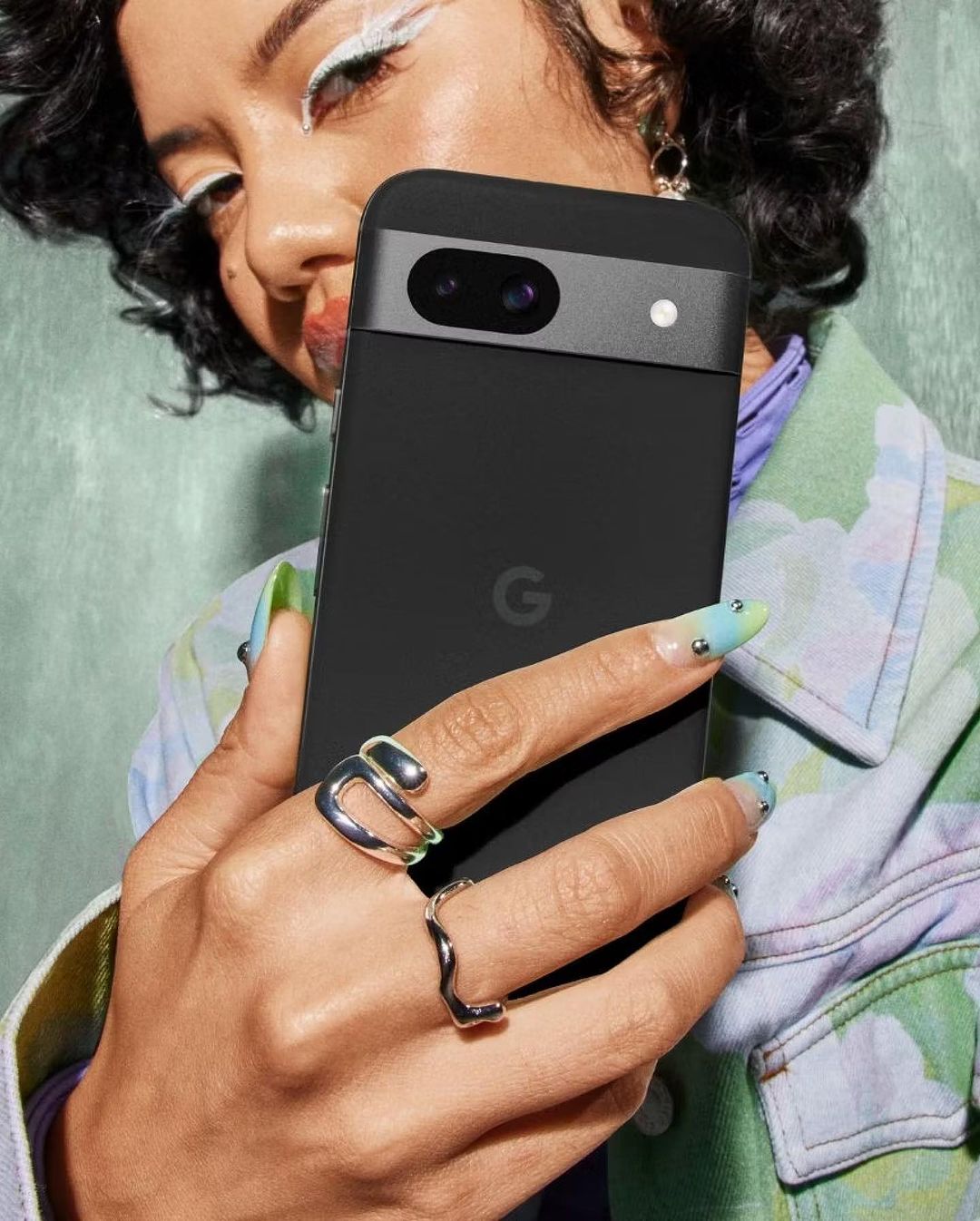 A woman with colorful eye makeup holding a black Google Pixel phone, showcasing silver rings and artistic nail designs against a green backdrop.