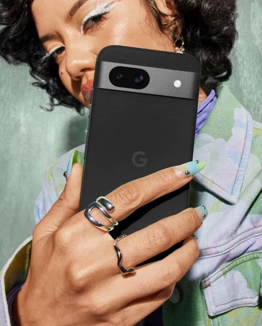 A woman with colorful eye makeup holding a black Google Pixel phone, showcasing silver rings and artistic nail designs against a green backdrop.