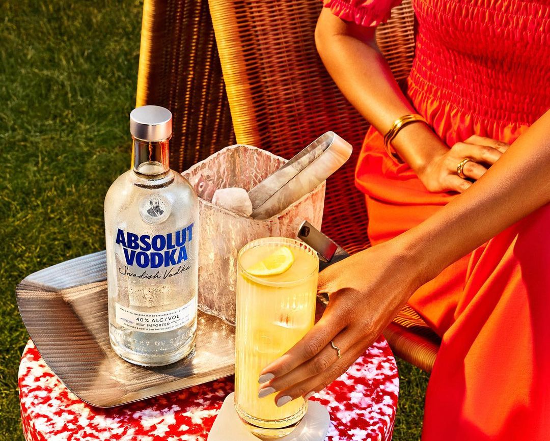 Woman in orange dress reaches for cocktail next to Absolut Vodka bottle, ice bucket, on red-and-white table in outdoor setting.