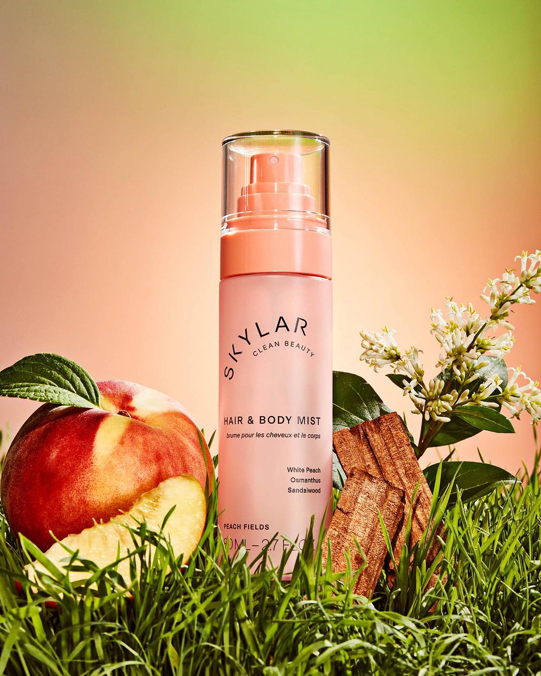 Skylar Clean Beauty hair and body mist bottle alongside a sliced peach, greenery, and floral accents against a gradient backdrop.