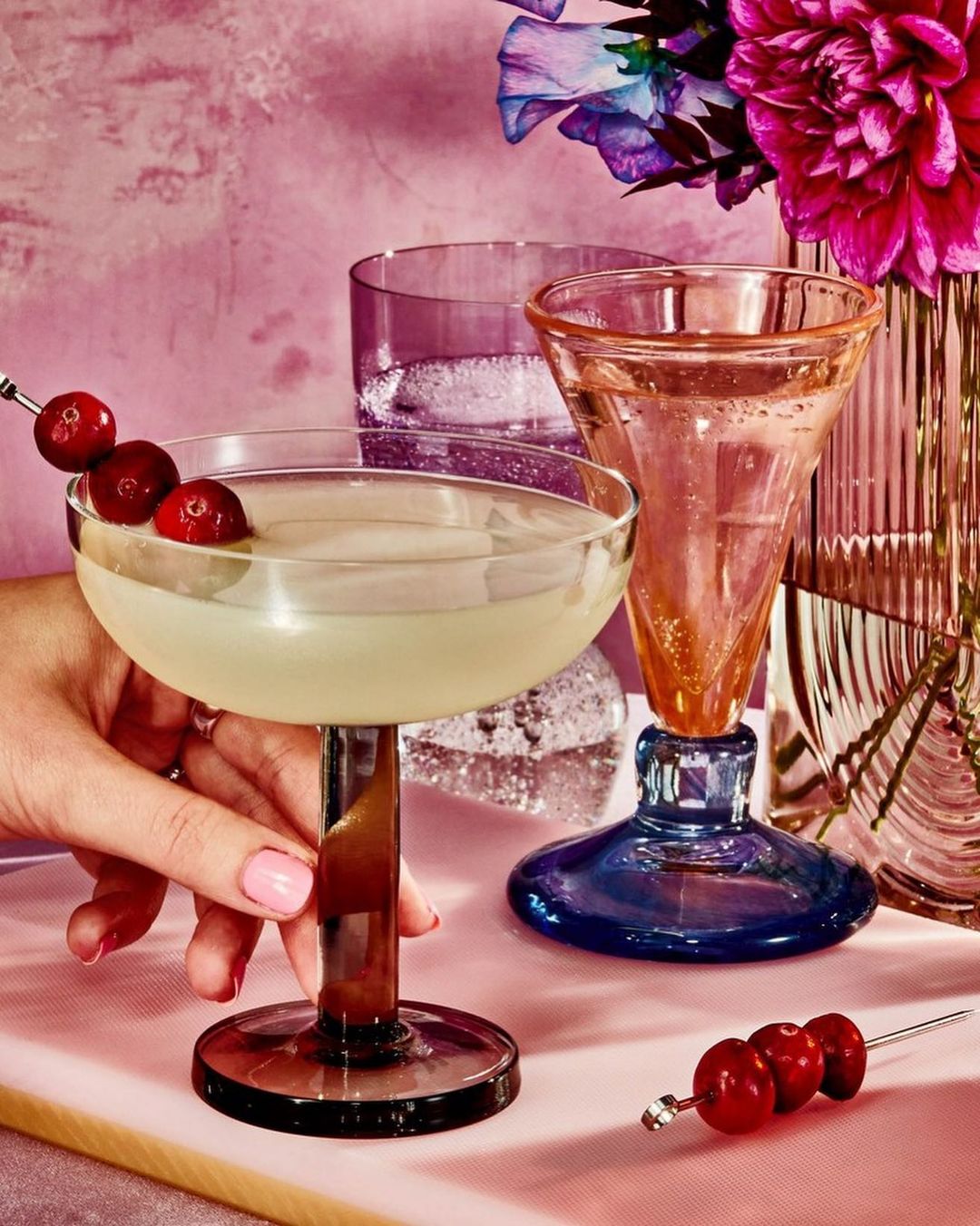 Close-up of a hand holding a cocktail glass filled with a drink and garnished with cranberries, with colorful glassware and flowers.