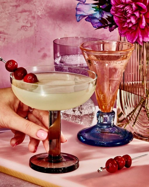 Close-up of a hand holding a cocktail glass filled with a drink and garnished with cranberries, with colorful glassware and flowers.