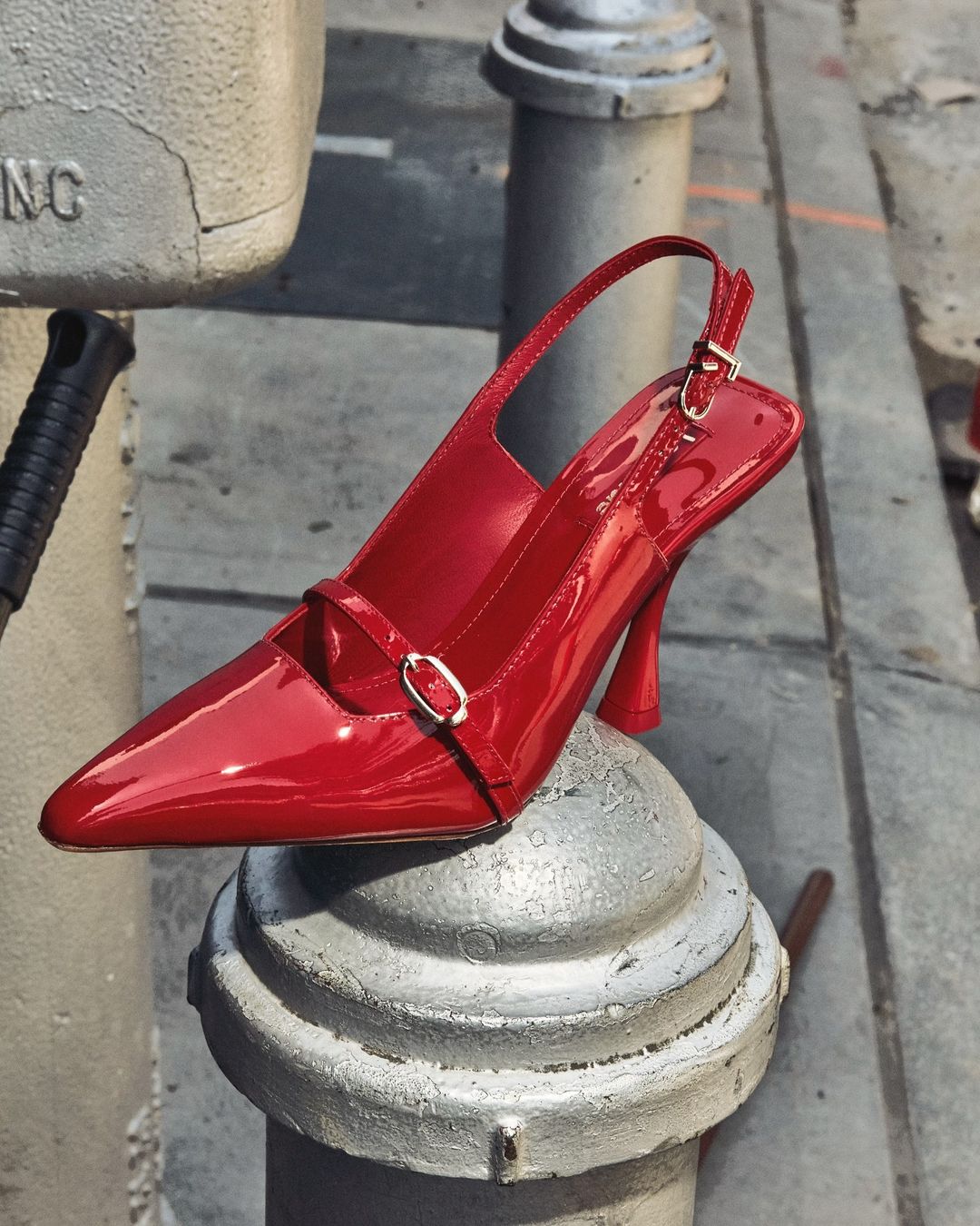 Red slingback high heel shoe displayed on top of a gray metal bollard in an outdoor urban setting, fashion product shot.