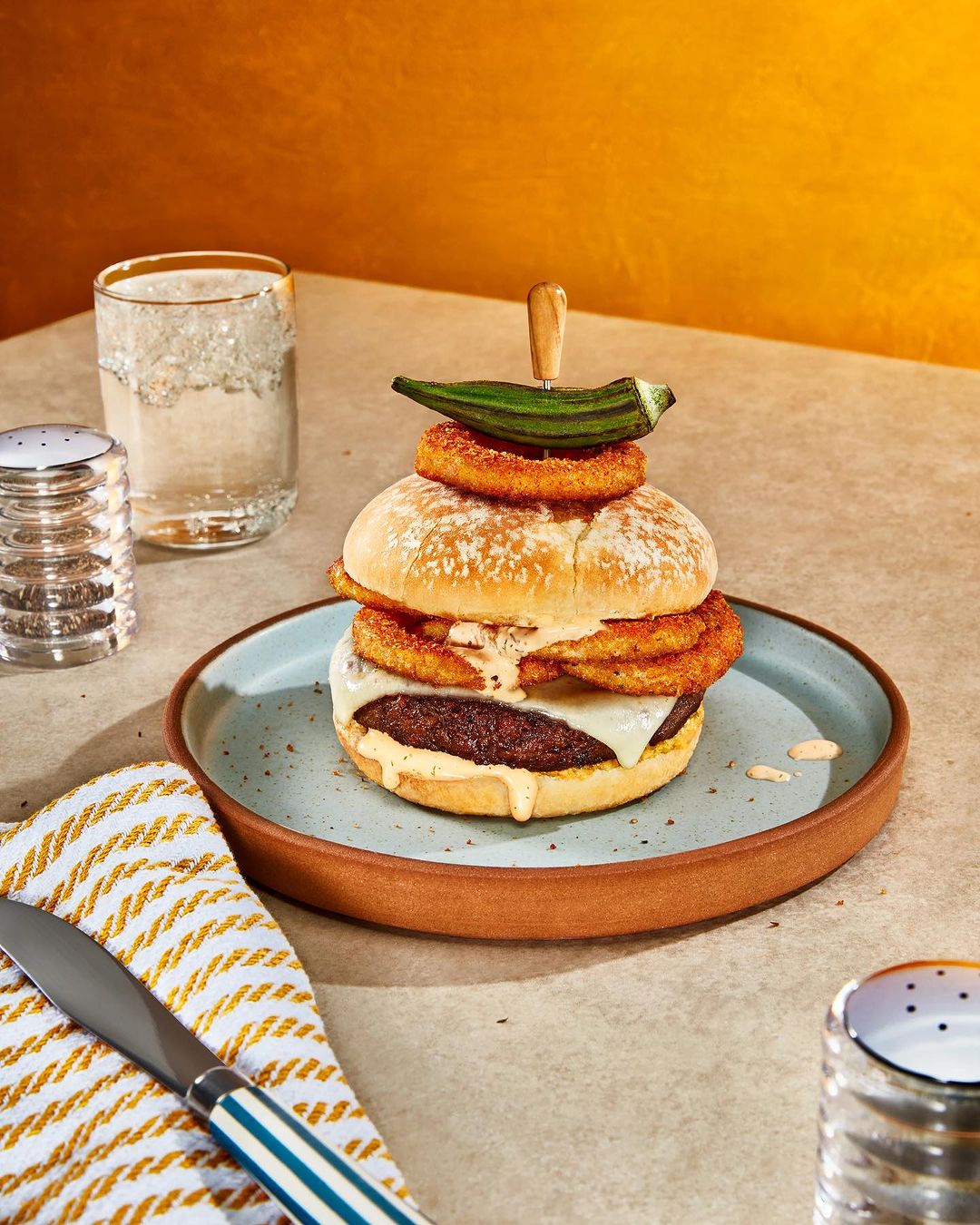 Gourmet burger with onion rings and okra garnish on a plate, accompanied by a knife and condiments against a warm backdrop.