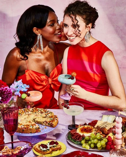 Two women in red dresses share a table of food and drinks. One woman whispers in the other's ear, both smiling with decorative jewelry.
