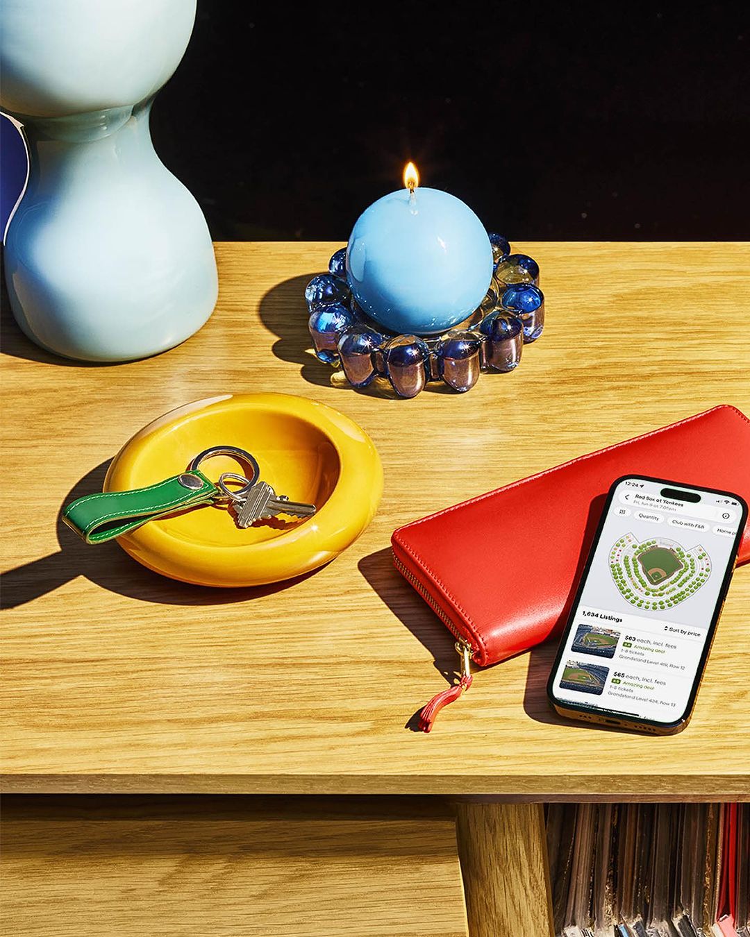 Still life featuring a lit blue candle, keys in a yellow dish, red wallet, and a smartphone displaying a stadium seating map on a wooden table.