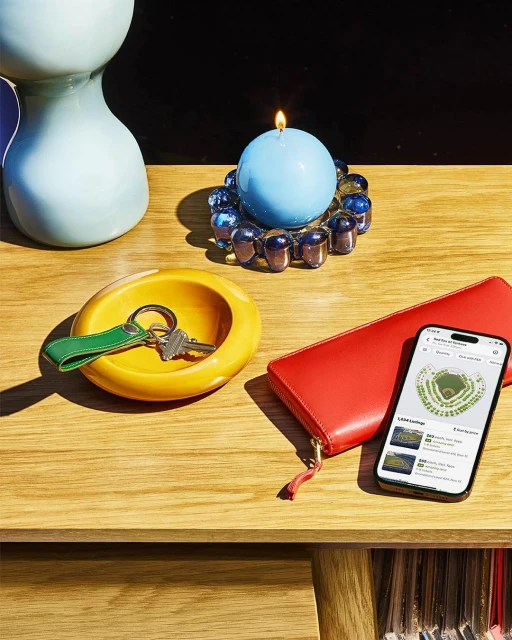 Still life featuring a lit blue candle, keys in a yellow dish, red wallet, and a smartphone displaying a stadium seating map on a wooden table.