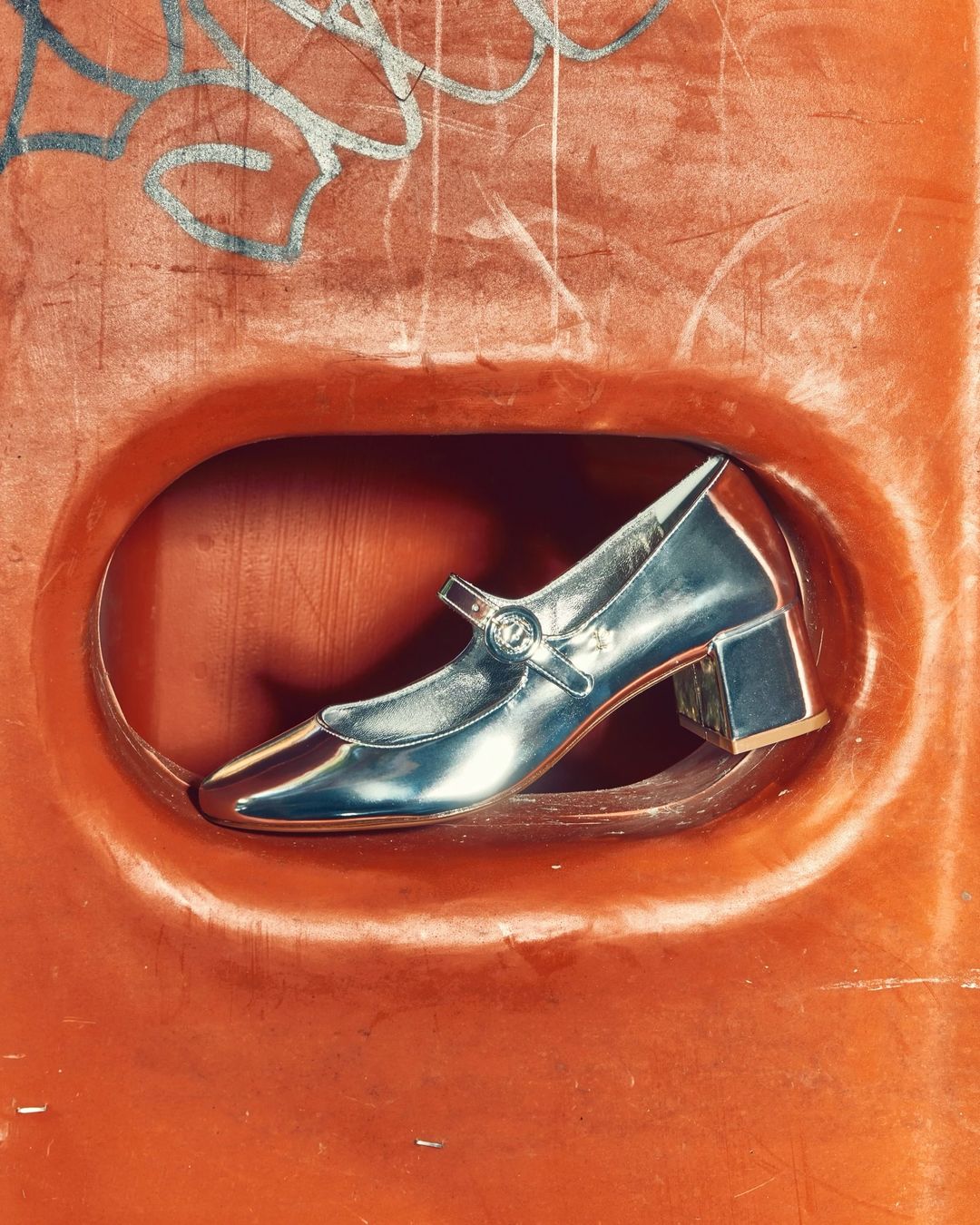 Elegant silver Mary Jane heel pump shoe with a block heel set against a textured orange-painted background with subtle graffiti detail.