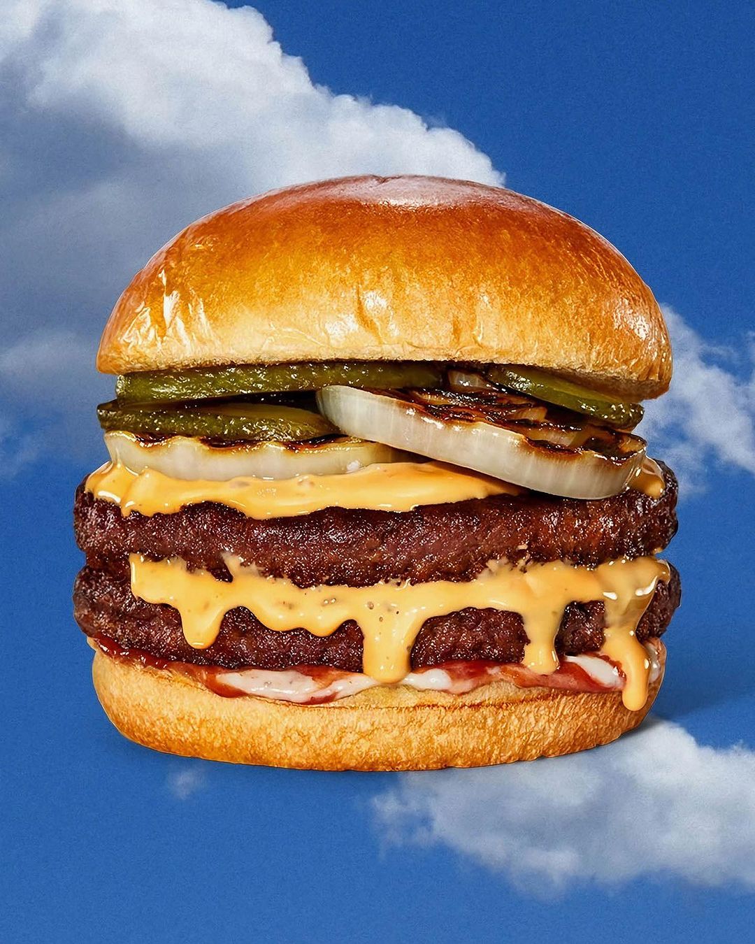 Double cheeseburger featuring beef patties, cheese sauce, grilled onions, and pickles on a bun, set against a blue sky background.