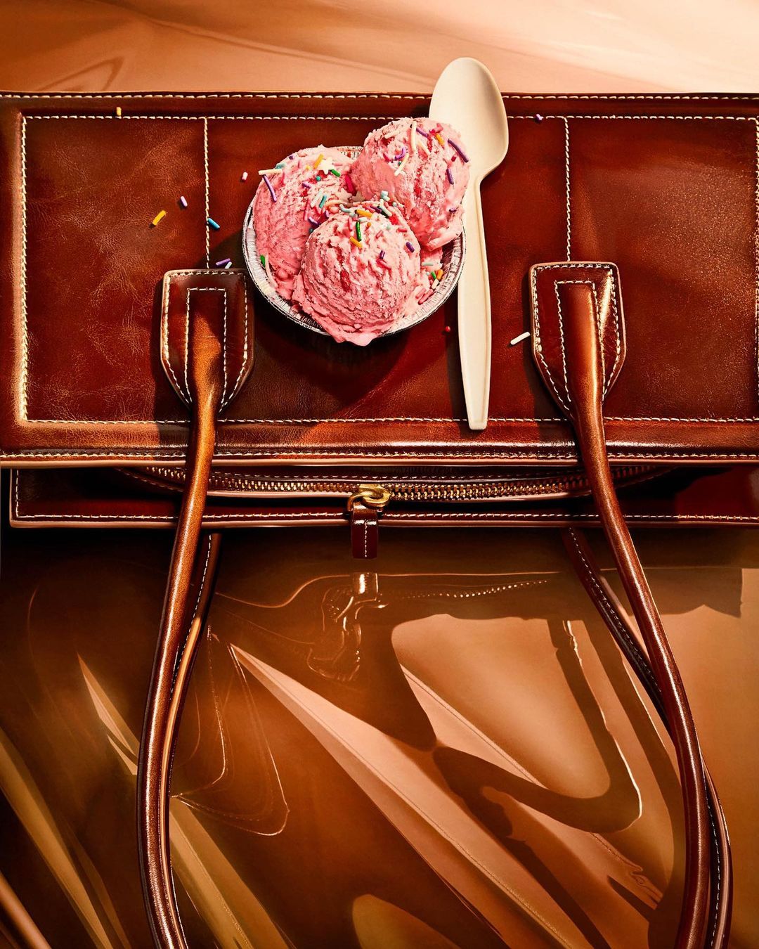 Overhead shot of pink ice cream with sprinkles in a bowl with a spoon, resting on a brown leather purse.