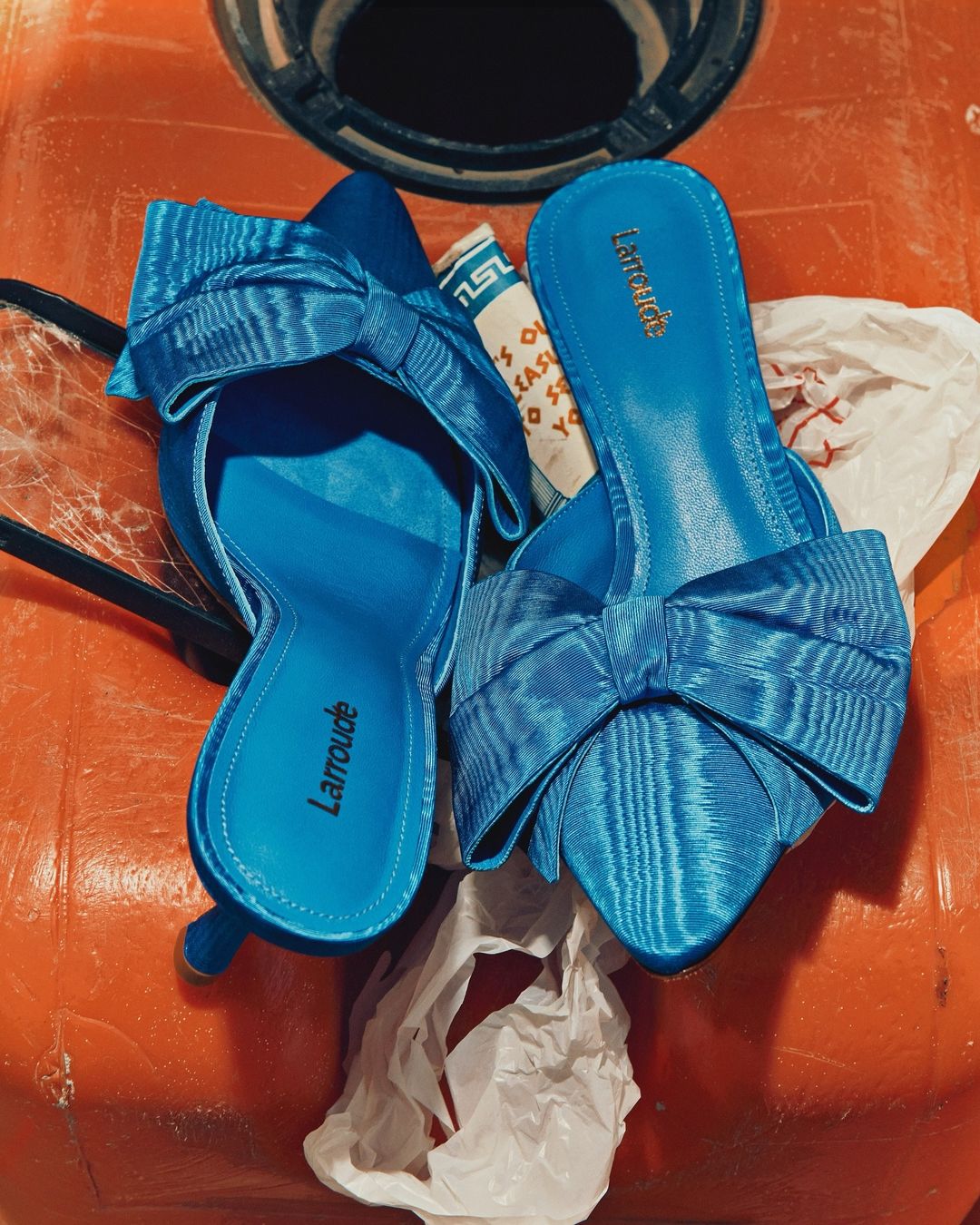 Close-up of bright blue heeled mules with bow detail, featuring the Larroude logo, with hints of an orange background.