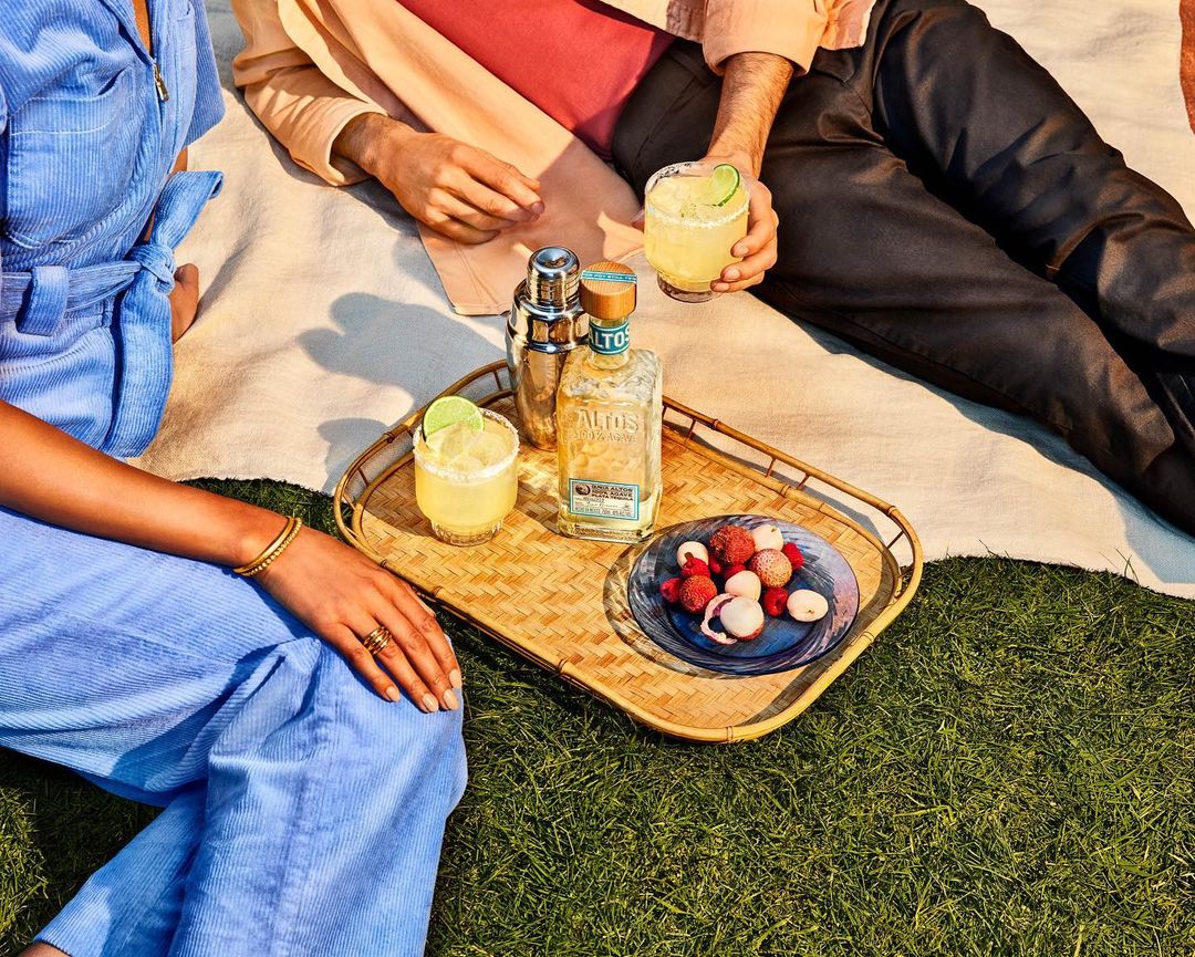 Two people sitting on a blanket outdoors with cocktails, liquor bottle, metal shaker, and a plate of fruit on a wicker tray.
