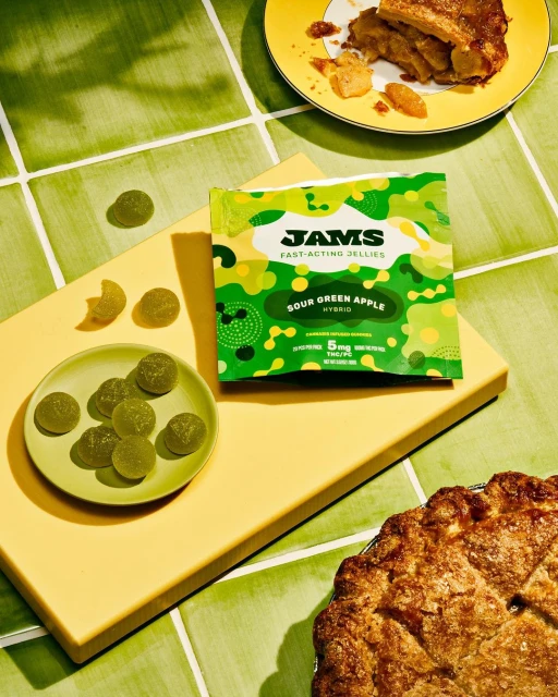 A bright overhead shot displays JAMS Sour Green Apple jellies with a slice of apple pie on a yellow and green tile backdrop.
