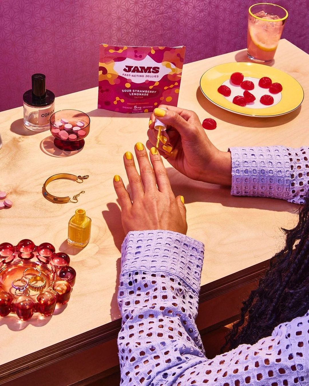 Woman applies yellow nail polish at table with gummies, drink, and beauty products in a brightly-lit and colorful indoor setting.