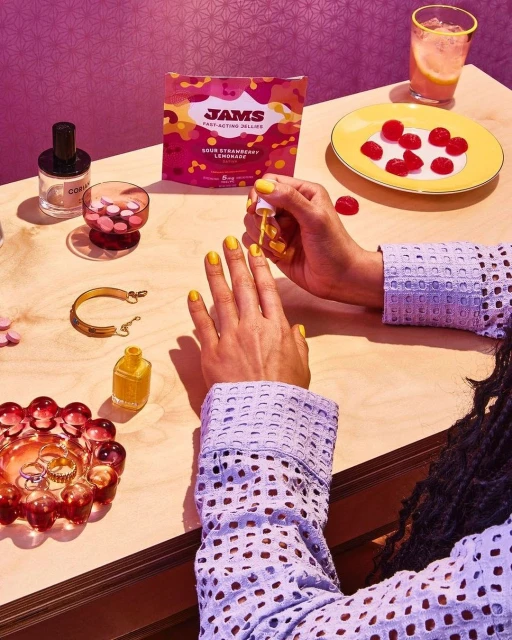 Woman applies yellow nail polish at table with gummies, drink, and beauty products in a brightly-lit and colorful indoor setting.