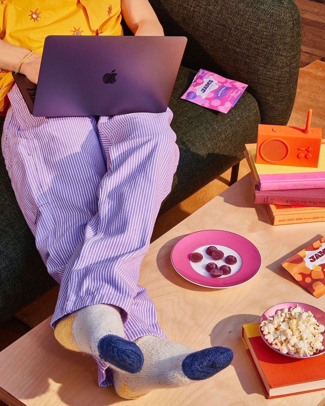 Person working on laptop, wearing striped pyjama pants and colourful socks. Snacks and a vintage-style radio sit on a wooden table.