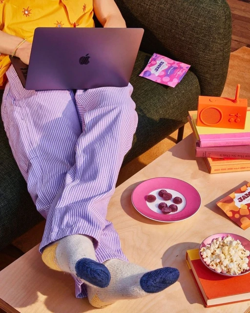 Person working on laptop, wearing striped pyjama pants and colourful socks. Snacks and a vintage-style radio sit on a wooden table.