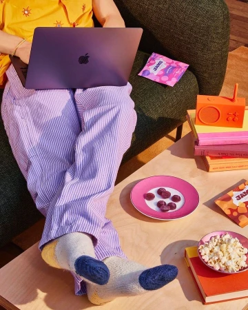 Person working on laptop, wearing striped pyjama pants and colourful socks. Snacks and a vintage-style radio sit on a wooden table.