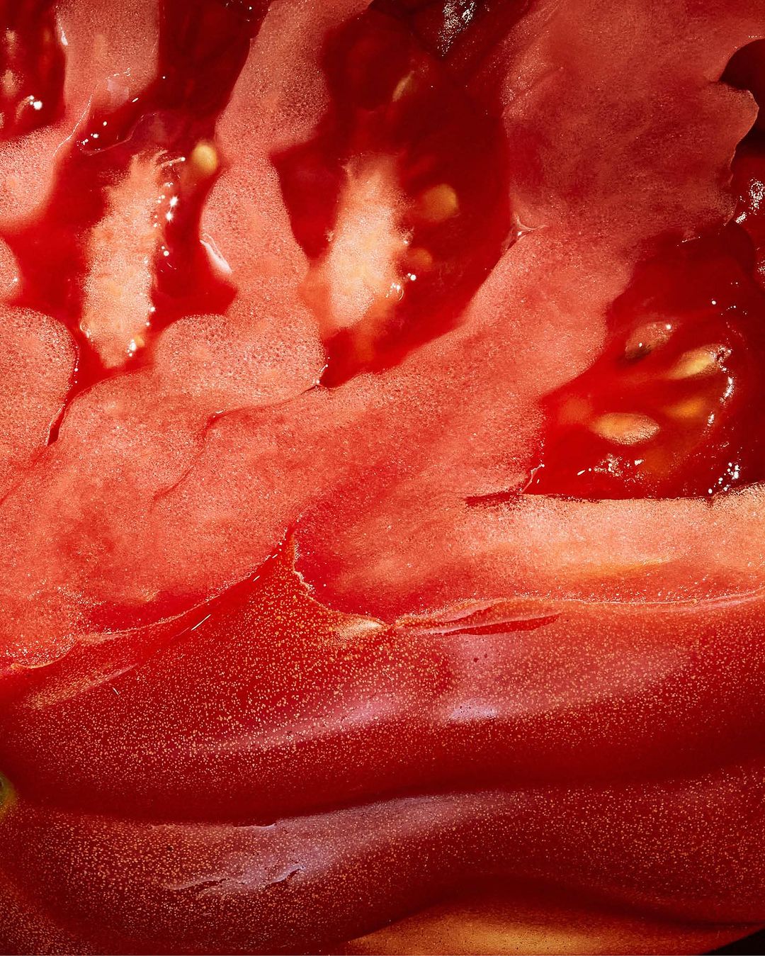 Close-up photograph of a sliced red tomato revealing its juicy interior, seeds, and fleshy texture, creating an abstract display of food.