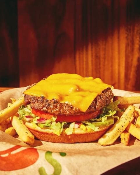 Overhead view of a cheeseburger with lettuce, tomato, onion, pickles, and fries on a branded paper liner against a wood background.