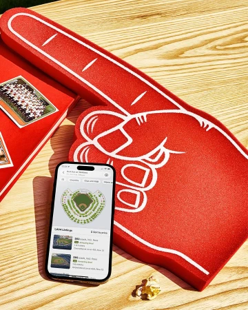 Overhead shot of an iPhone displaying a stadium seating chart next to a foam hand and a book on a wooden table.