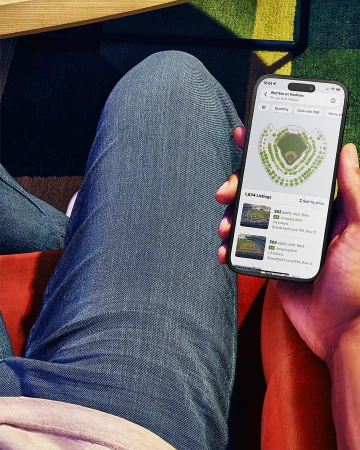 Person holding a smartphone displaying a stadium seating chart application, knees and legs visible in the frame.