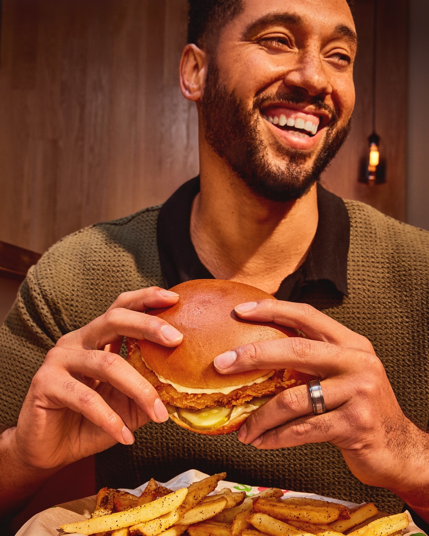 Smiling man holds chicken sandwich over plate of fries. Dark hair, ring, and olive green top are visible in an indoor setting.