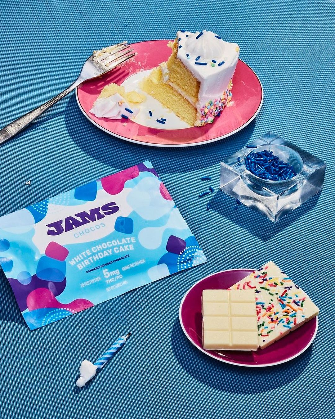 Overhead shot of a dessert table with a slice of cake, chocolate bars, a birthday candle, and sprinkles against a blue surface.