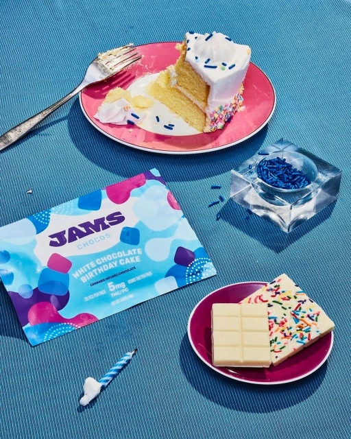 Overhead shot of a dessert table with a slice of cake, chocolate bars, a birthday candle, and sprinkles against a blue surface.