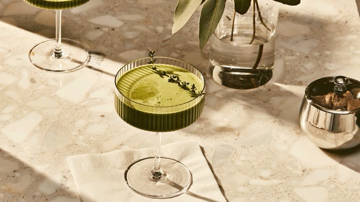 Two green cocktails served in coupe glasses on a marble table, garnished with sprigs of thyme, next to a vase of greenery.