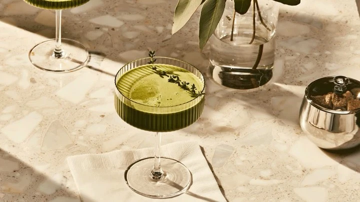 Two green cocktails served in coupe glasses on a marble table, garnished with sprigs of thyme, next to a vase of greenery.