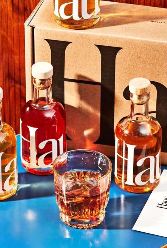 Still life arrangement of Haus aperitif bottles and a glass of cocktail with ice, placed on a blue surface in a studio setting.