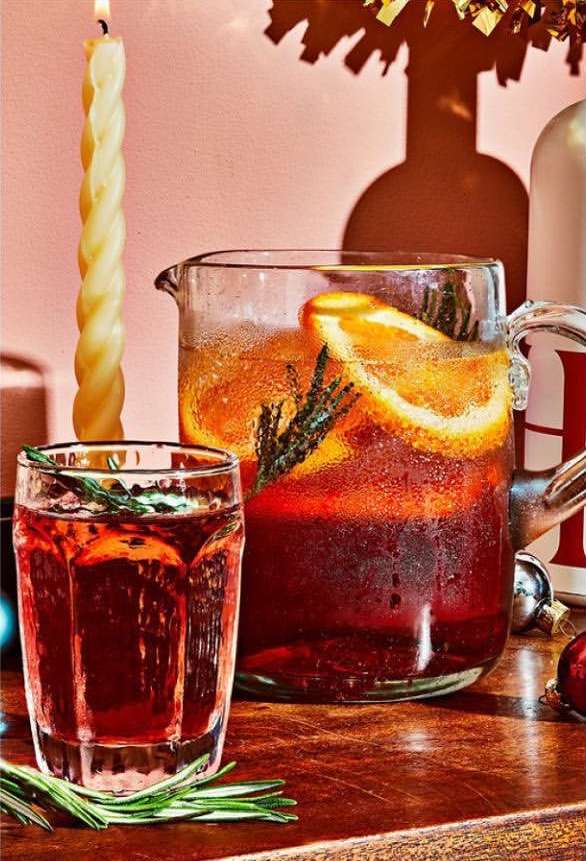 Festive drink in glass and pitcher with orange slices and rosemary sprigs, with a lit candle, on a wooden surface.