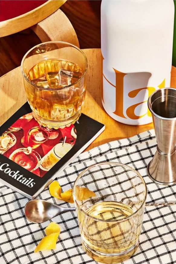 Two cocktails with ice, a jigger, and lemon peel sit on a black and white checkered cloth and wooden surface, in a warm, inviting setting.