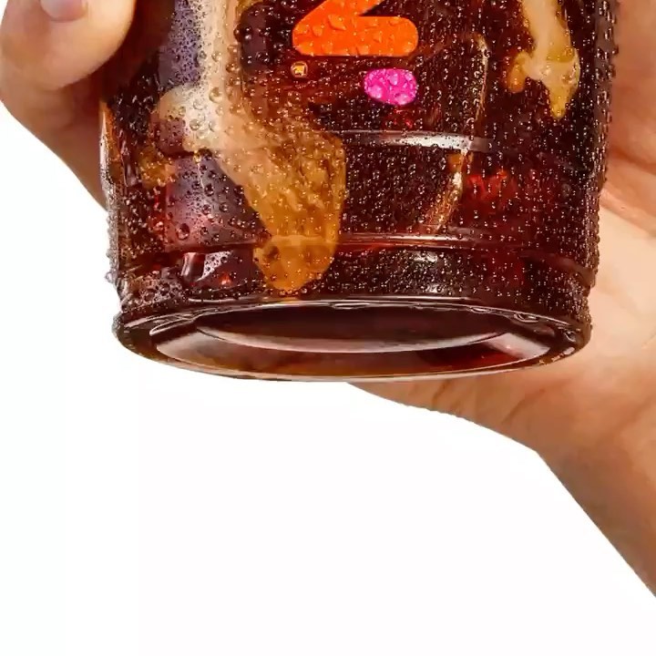 Close-up of a clear plastic cup with a brown beverage and condensation, held by a hand against a white background.