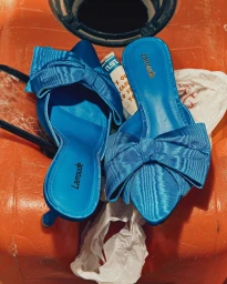Top-down view of vibrant blue satin mules with prominent bows resting on a bright orange surface.