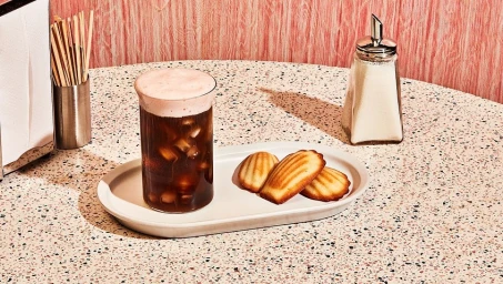 Iced coffee in a ribbed glass with three madeleines on a white tray on a speckled tabletop with a sugar dispenser