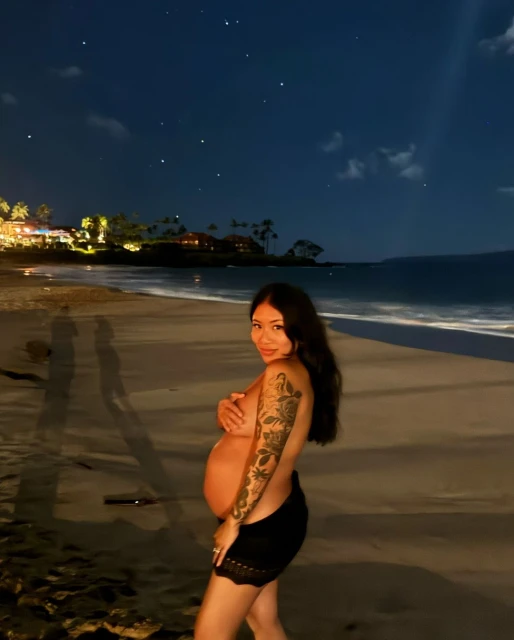 Pregnant Asian woman poses at night on a sandy beach with illuminated buildings and palm trees under a starry, moonlit sky.