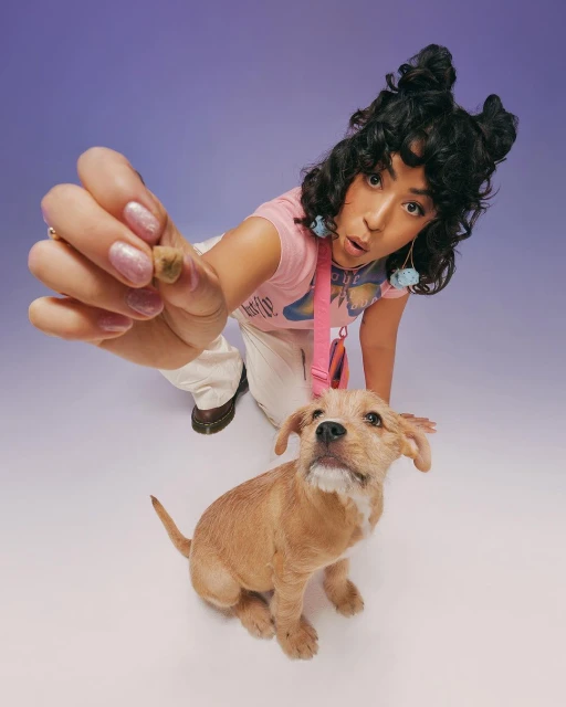 Young woman crouching with her dog as she holds out a treat indoors in front of a purple studio backdrop with bright lighting.