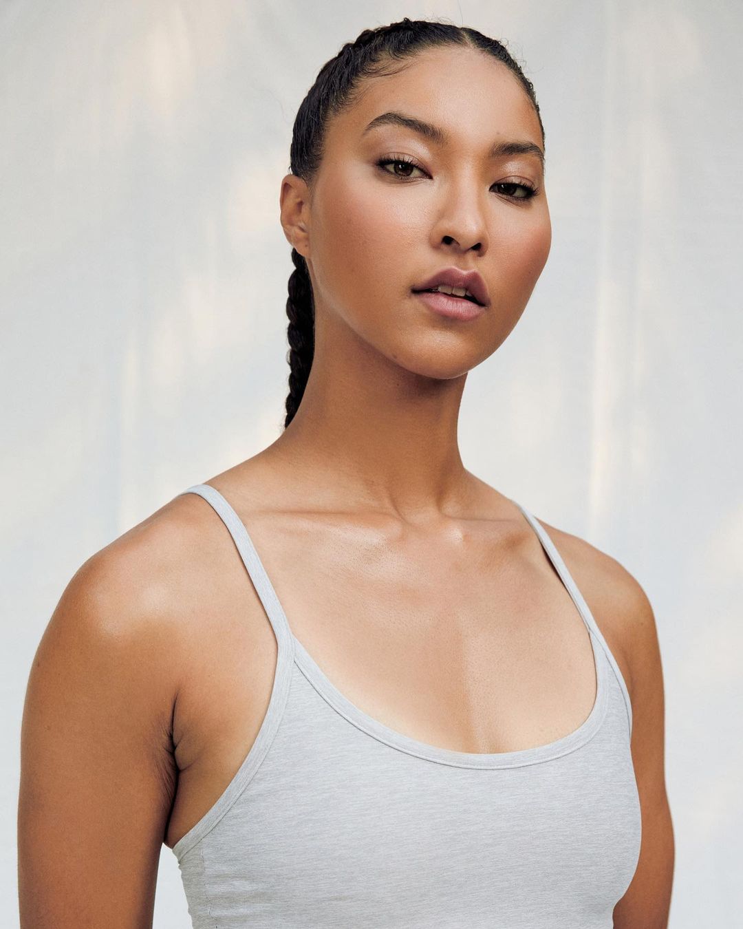 Close-up portrait of a young adult female with braided hair and a grey top against a bright, diffused white background.