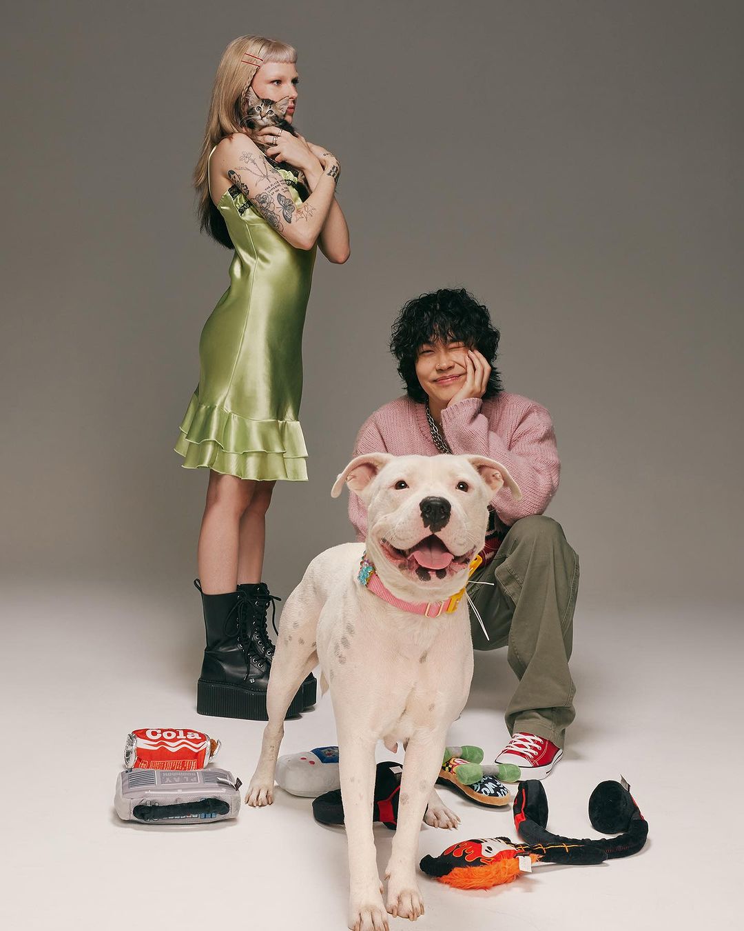 Two people pose with a white pitbull dog in a studio setting; a woman stands in a green dress with a man kneeling with the dog.