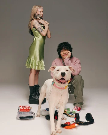 Two people pose with a white pitbull dog in a studio setting; a woman stands in a green dress with a man kneeling with the dog.