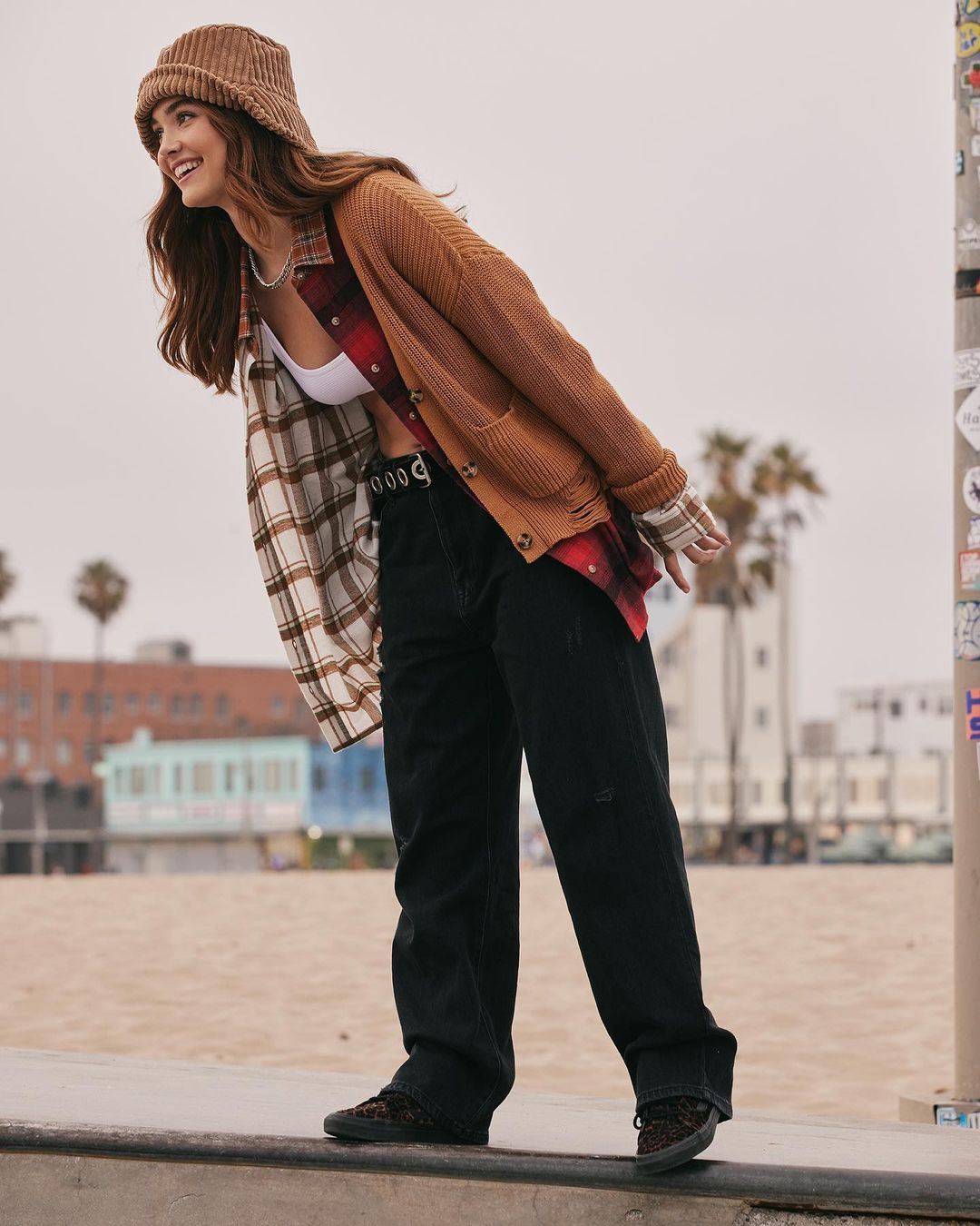 A young woman in a cardigan and bucket hat poses outdoors on a concrete barrier near a beach and building with palm trees in the background.