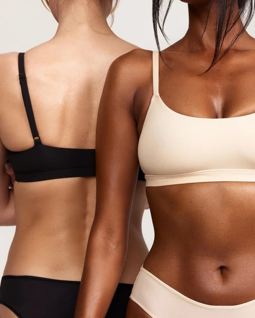 Close-up studio shot of two adult women, one with light skin and one with dark skin, posing in matching neutral-tone underwear sets.