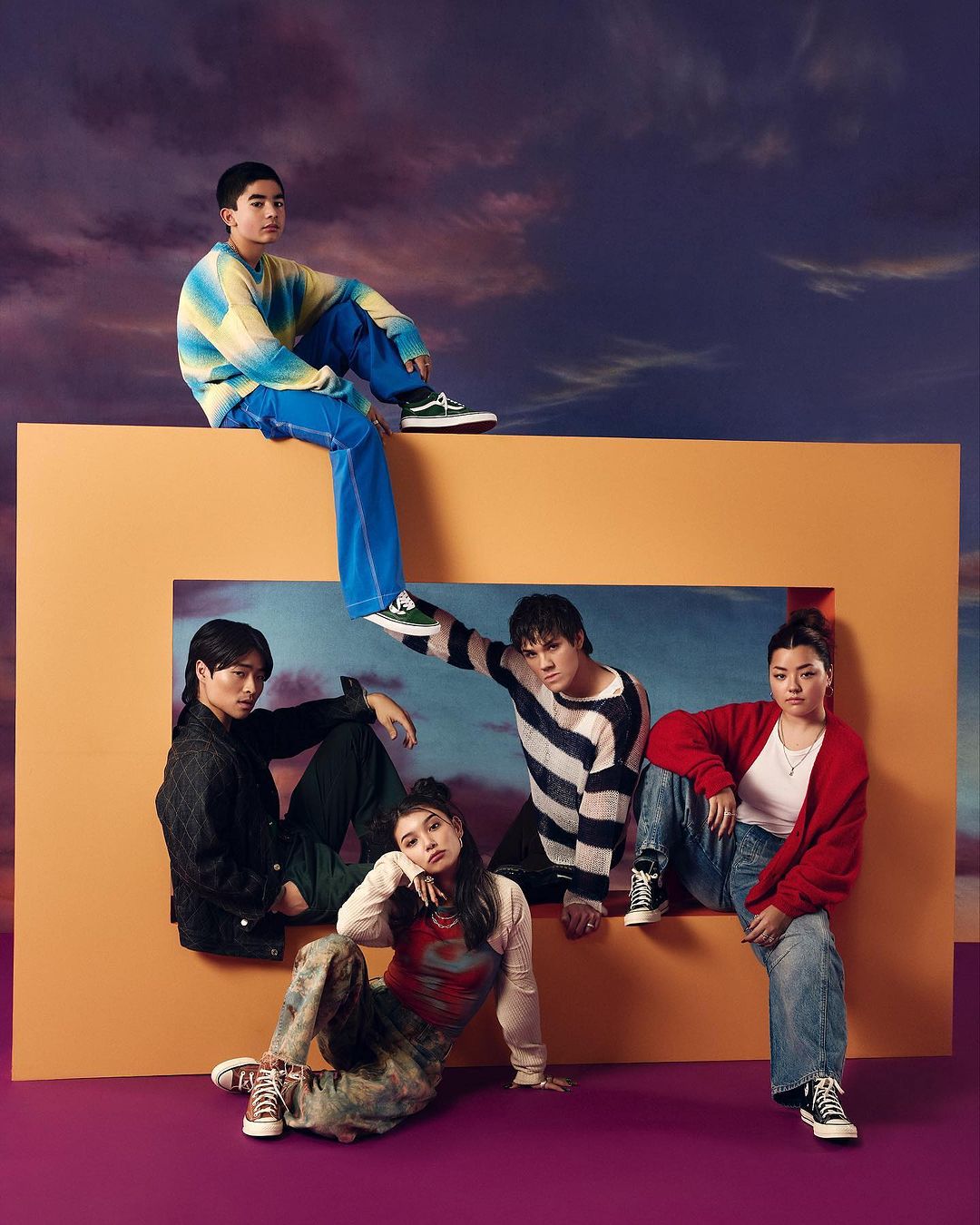 Group of five diverse young adults posing together in a studio with orange frame prop against a sky background, wearing casual clothing.