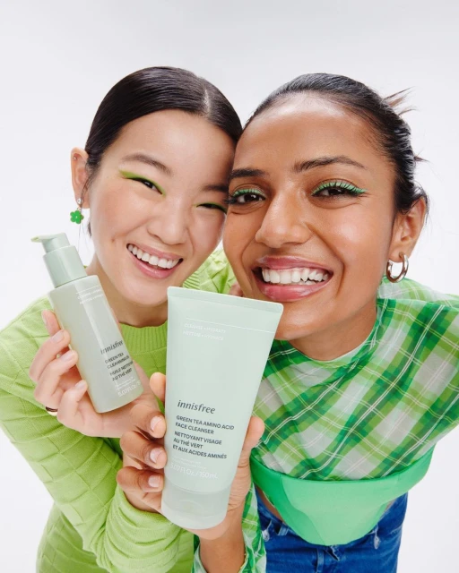 Two smiling women holding skincare products. One holds a cleanser tube, and the other displays a serum bottle in a bright studio setting.
