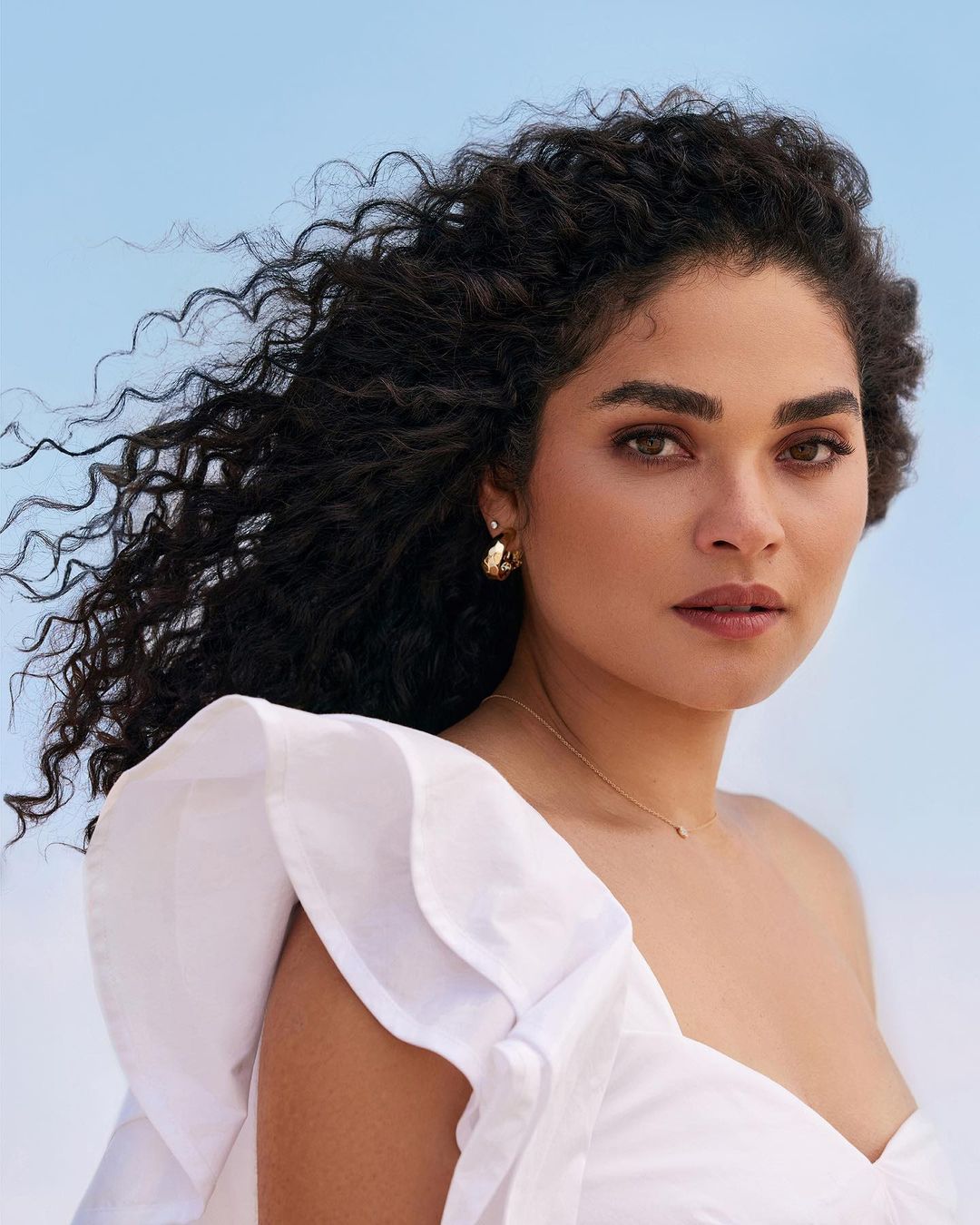 Close-up of a beautiful woman with curly hair wearing a white one-shoulder top against a light blue sky background, portrait orientation.